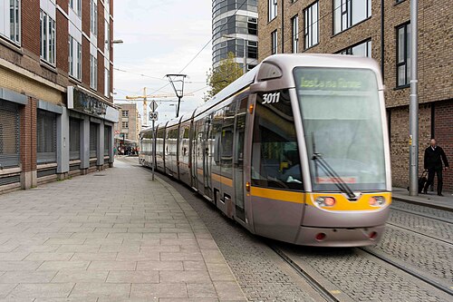A Luas tram running along the paved and cobbled street beside brick and glass apartment buildings.