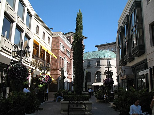 The cobblestone court of Two Rodeo Drive, a tall clipped cypress at the center, a brick building with yellow awnings and hanging flower baskets on the lampposts.
