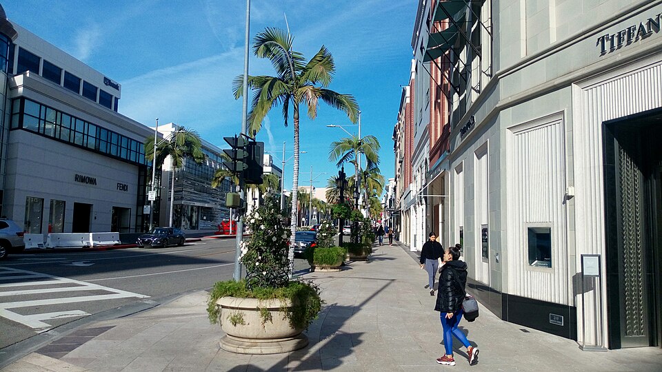 Rodeo Drive's sidewalk, storefronts for Tiffany, Rimowa and Fendi, palm trees down the median and a woman walking past with shopping bags.