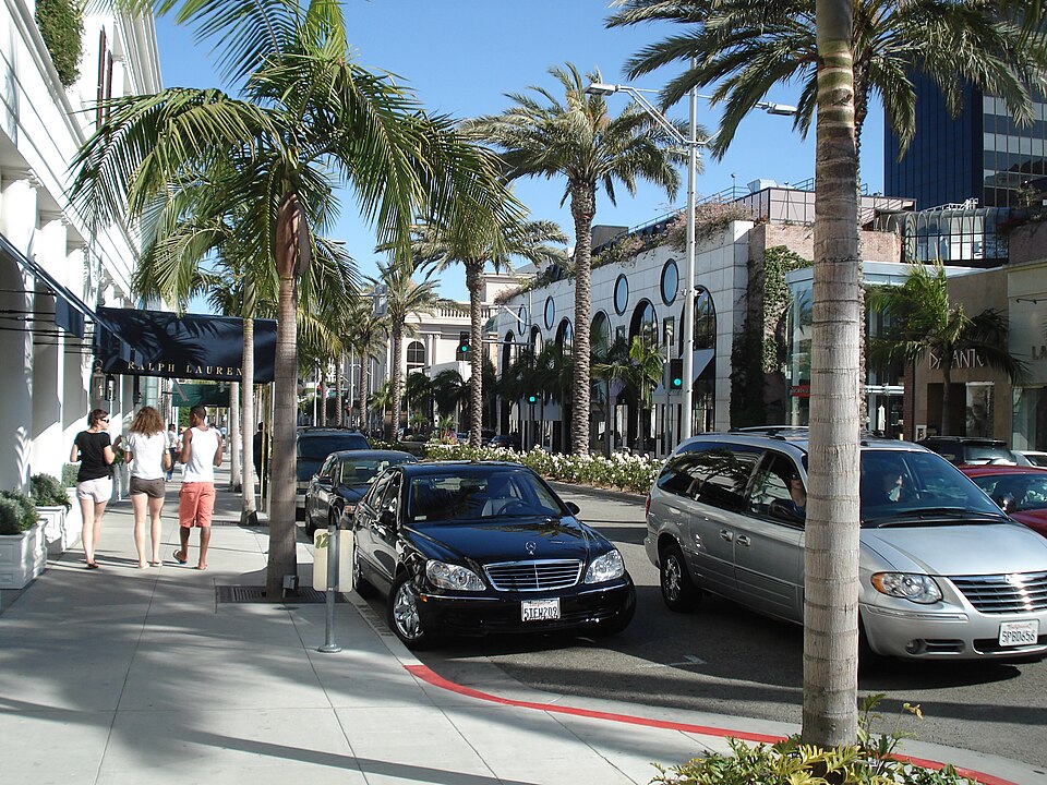 Rodeo Drive under a row of palm trees, a Ralph Lauren awning on the near corner, cars angled into the curb and a rounded white building with porthole windows down the block.