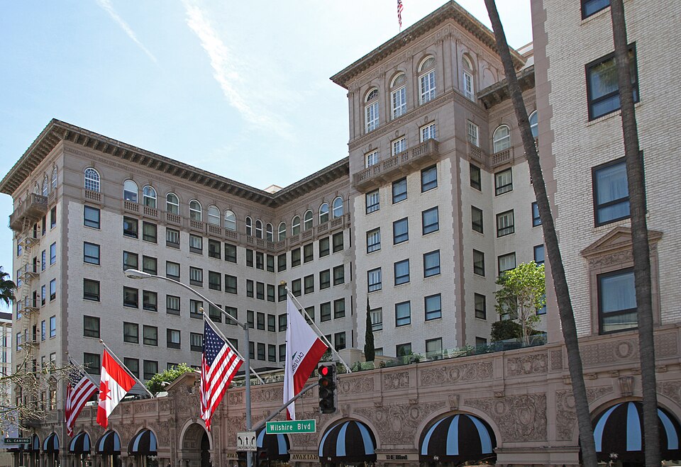 The Beverly Wilshire Hotel's corner at Wilshire Boulevard, American, Canadian and California flags out front, striped awnings over the entrances and palm trees at the curb.