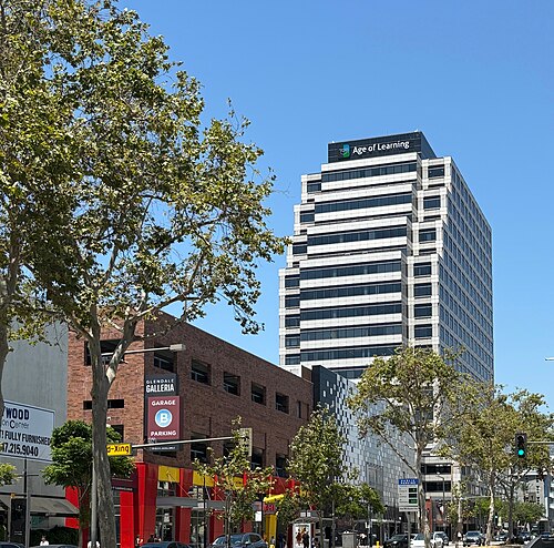 Age of Learning's office tower behind the Glendale Galleria's parking garage sign, palm trees and storefronts along the sidewalk.