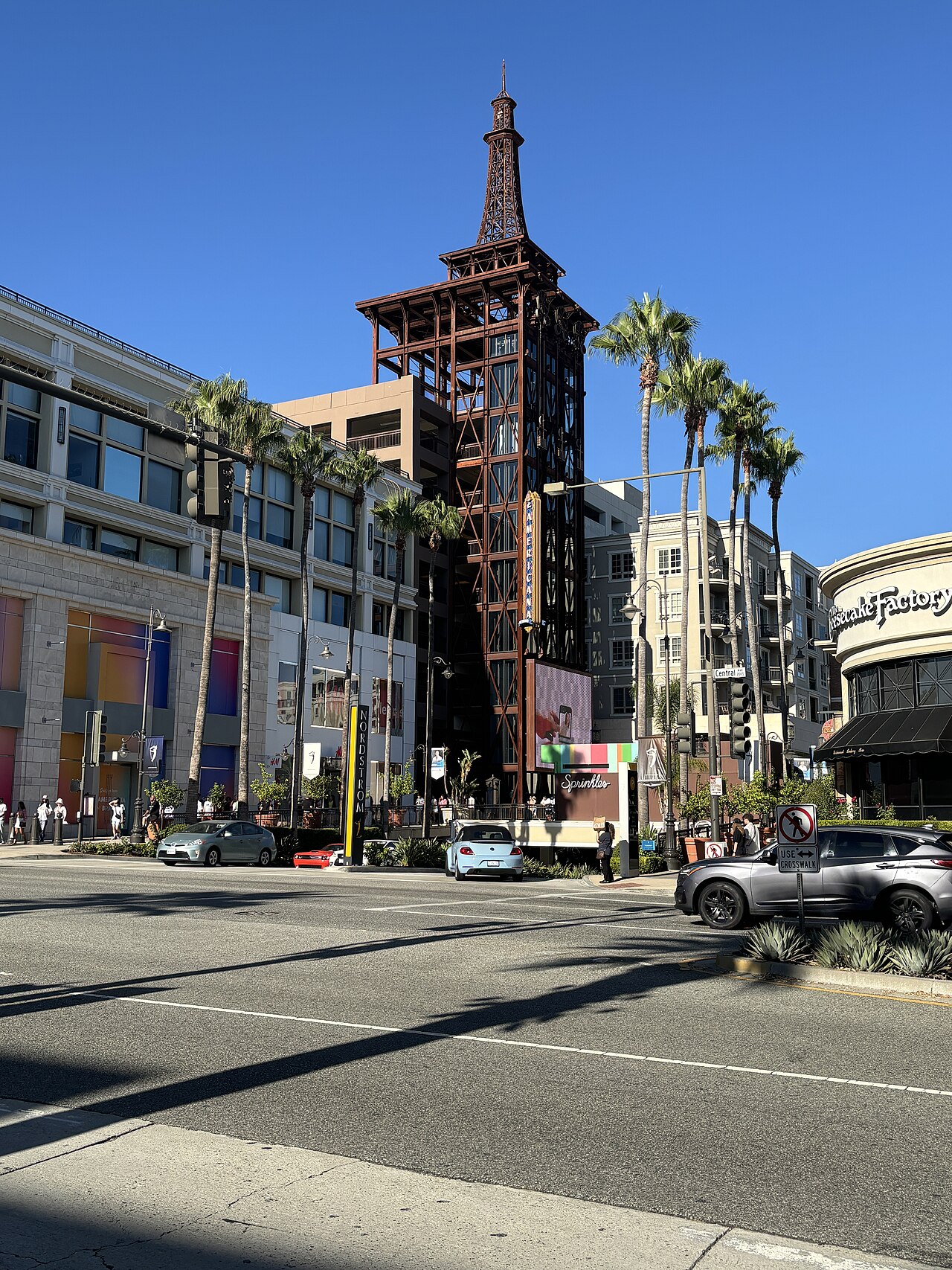 The Americana at Brand's steel elevator tower rising over the plaza on Brand Boulevard, palm trees and storefronts on both sides, Nordstrom to the left and the Cheesecake Factory to the right.