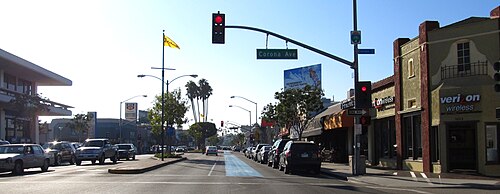Second Street at Corona Avenue, a Verizon Wireless and a Rite Aid pharmacy among the storefronts, palm trees at the median and cars parked along the curb.