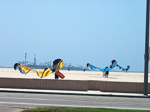 Kitesurfers laying out their kites on the sand along the boardwalk, palm trees behind them and a tower down the beach.