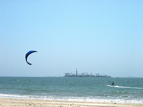 A kitesurfer working the water, with a small island carrying palm trees and a few buildings in the distance.