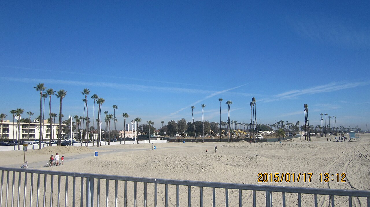 The beach seen from the Belmont Pier, palm trees and a beachfront apartment building on the left and grading equipment working the sand in the middle distance.