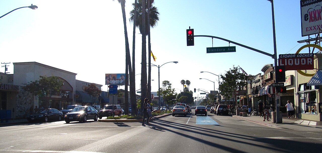 Second Street at Glendora Avenue, palm trees along the median, a liquor store and other storefronts on the right, cars stopped at a red light and a pedestrian crossing.
