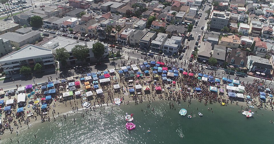 An aerial view of Horny Corner on the Fourth of July, tents and umbrellas lined along the sand at Alamitos Bay and houses on the street above the beach.