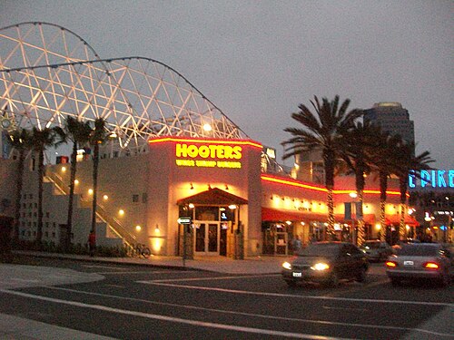 The Pike at Rainbow Harbor at night, restaurant signs and palm trees along the promenade, a lit steel arch shaped like a roller coaster behind them.