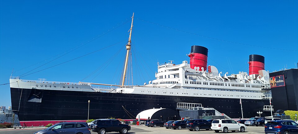 The RMS Queen Mary at her Long Beach berth, her black hull and red and black funnels against a clear sky, cars parked along the pier.