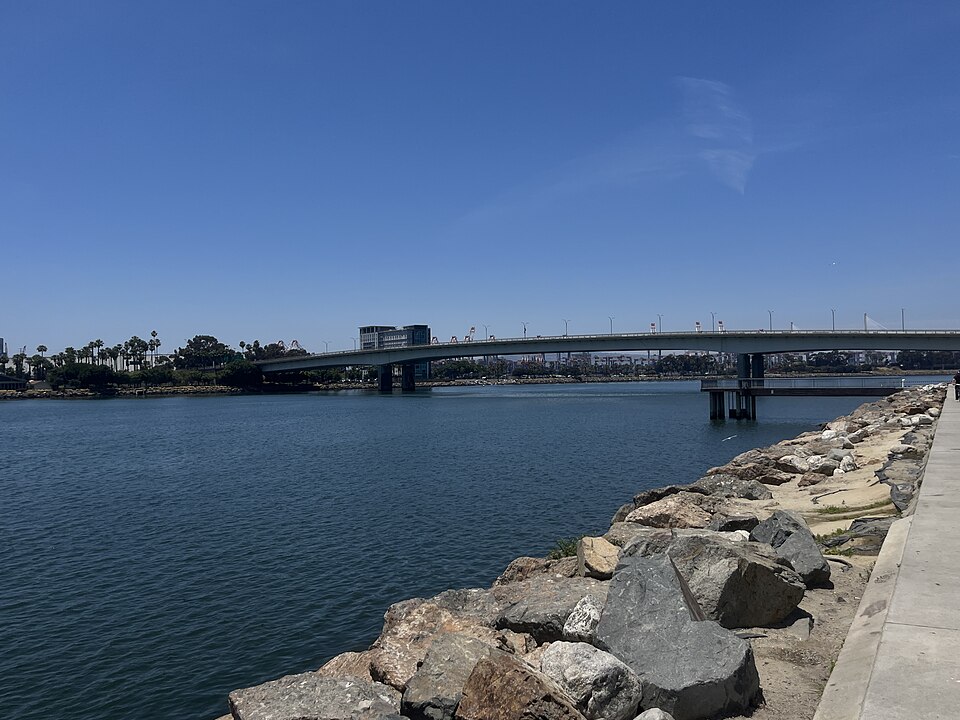 The Queensway Bridge seen across the back channel, a low concrete span carrying traffic between downtown and the harbor, a rock jetty in the foreground.