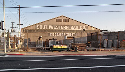 The Southwestern Bag Co. warehouse on East 6th Street, its name and address painted across the gable, drums and lumber stacked behind a razor wire fence.