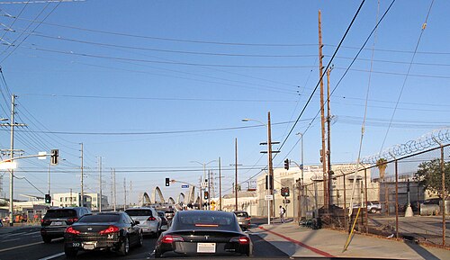 Sixth Street near Mateo, cars stopped at a light beside a chain link fence topped with razor wire, the arches of the Sixth Street Viaduct ahead.