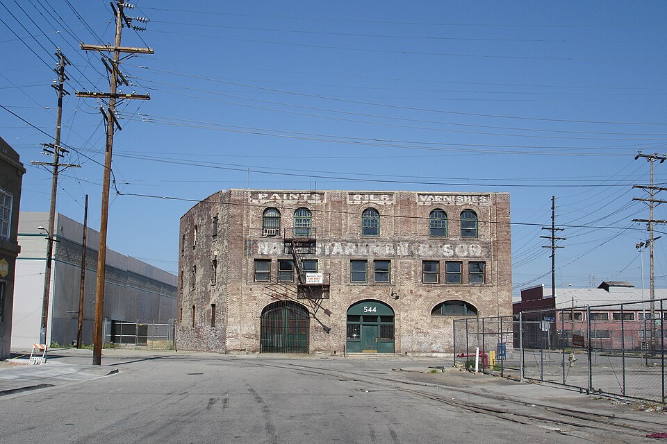 The Nate Starkman & Son warehouse at 544 Mateo Street seen across the empty intersection, PAINTS OILS VARNISHES and the company's name still legible in ghost lettering above the windows.