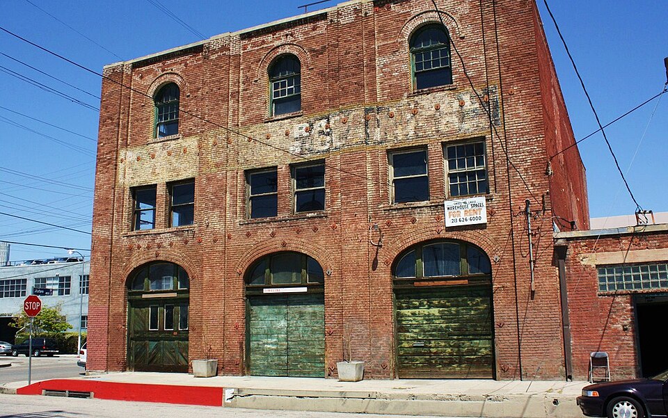 The same warehouse's corner entrance at street level, arched doorways with green doors, a red curb, and a warehouse space for rent sign over one window.