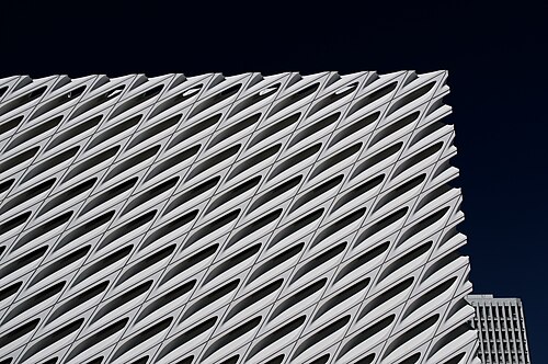 A corner of The Broad's white honeycomb exterior against a dark sky, the diagonal lattice of oculus-like openings running the length of the facade with a taller office tower visible behind it.