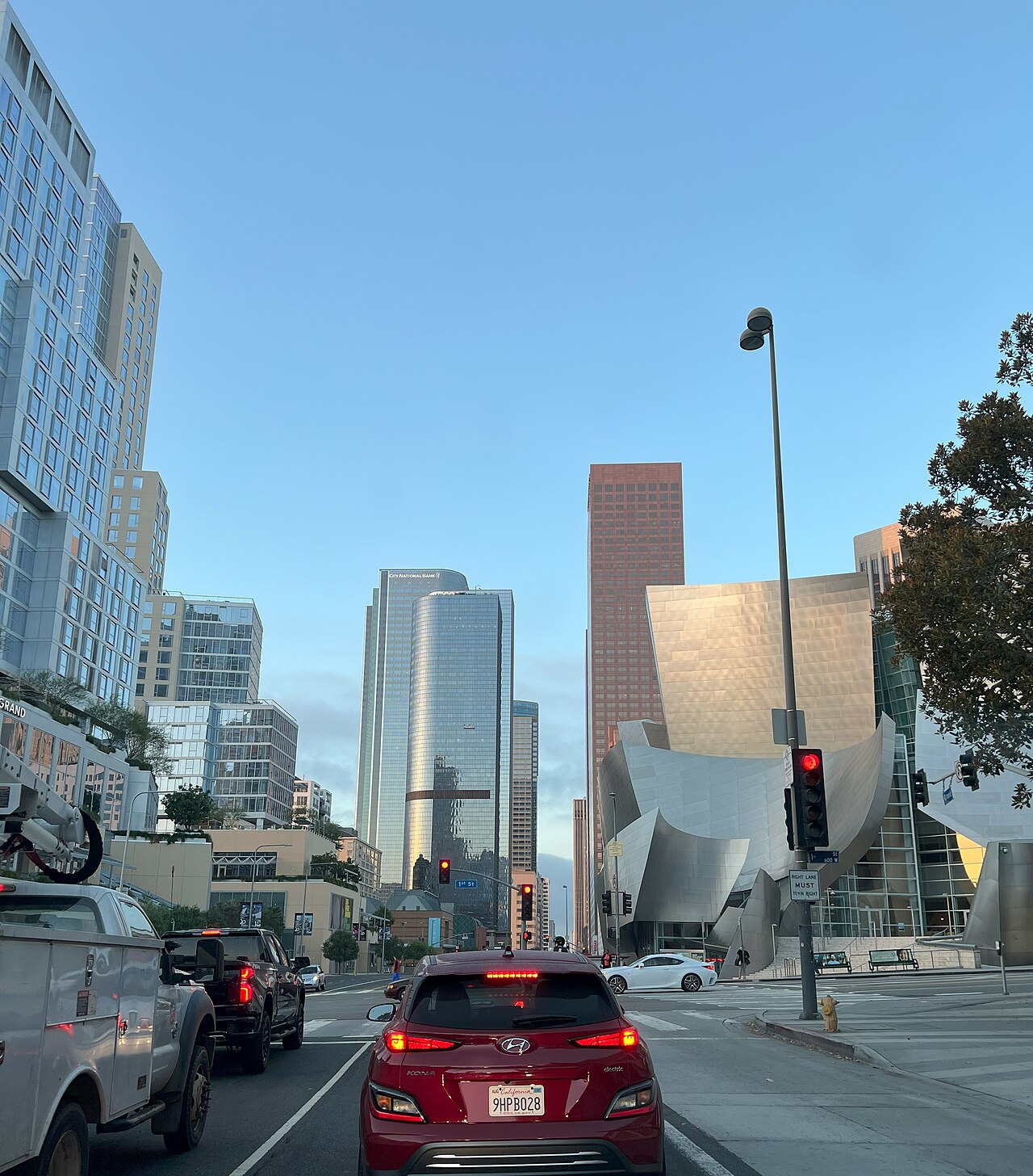 Grand Avenue looking south, the Walt Disney Concert Hall's steel curves catching the morning light on the right, more downtown towers filling the block behind it, and traffic stopped at a light in the foreground.
