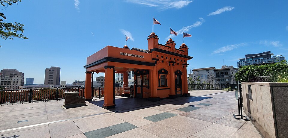 The orange upper station of the Angels Flight funicular, American flags on its towers, at the top of the incline with downtown towers behind it.
