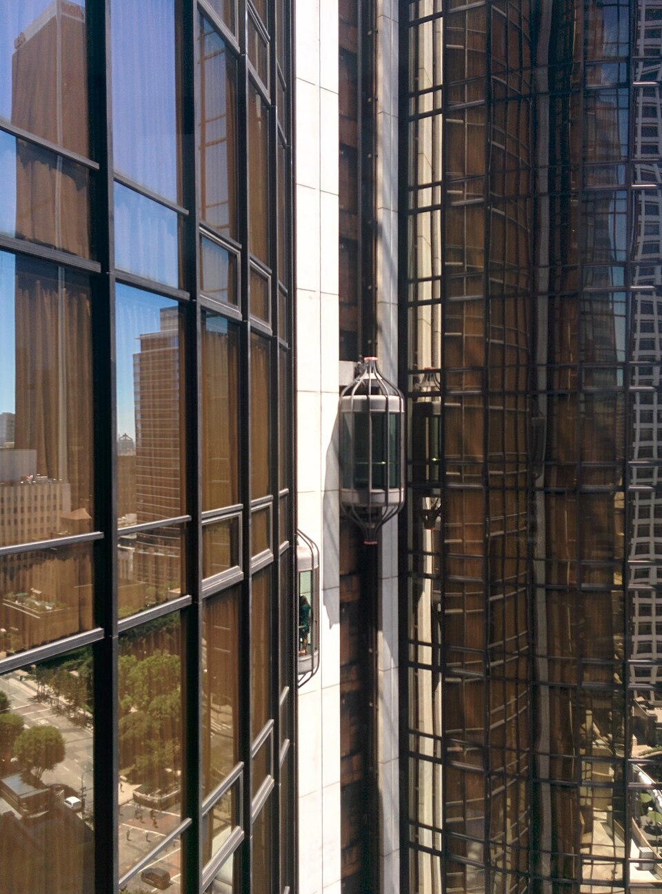 The Westin Bonaventure's external glass elevator pods running up a shaft between two of its mirrored cylindrical towers, downtown Los Angeles reflected in the glass.