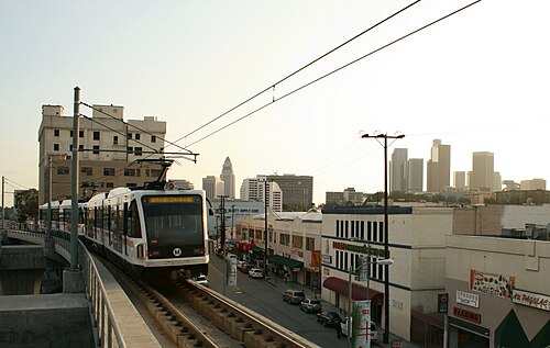A Metro Gold Line train on the elevated track above a Chinatown street, storefronts below and the downtown Los Angeles skyline behind.