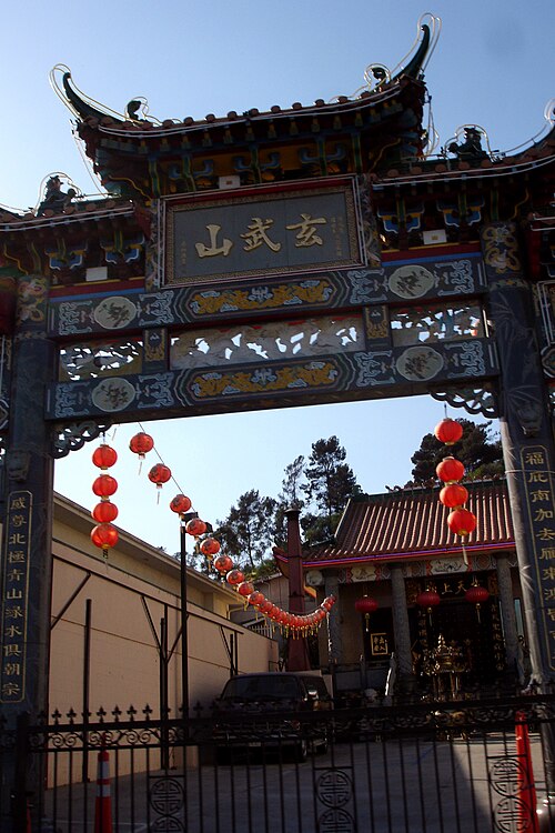 A paifang gate on Broadway, red lanterns hanging beneath its tiled roof, a temple building visible through the archway.