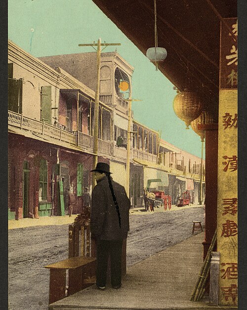 A street in Old Chinatown, paper lanterns hanging from an awning, a man standing at the curb and a horse drawn cart farther down the block.