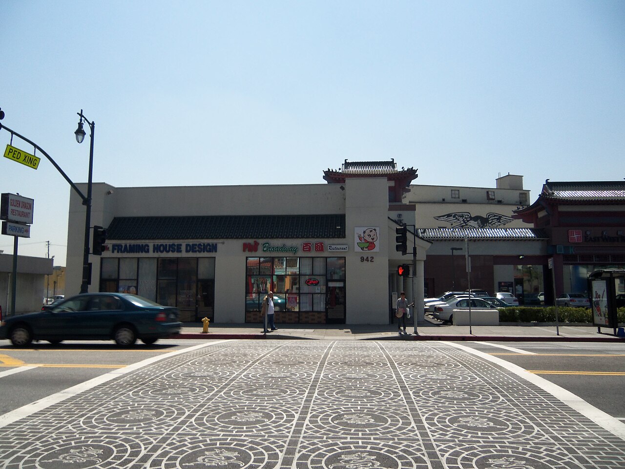 Broadway at 942 North Broadway, storefronts under a green awning, a pagoda roofed building beyond, and a crosswalk paved with dragon medallions.