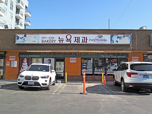 A bakery storefront with one sign lettered in English, Korean and Spanish, cars parked in front.