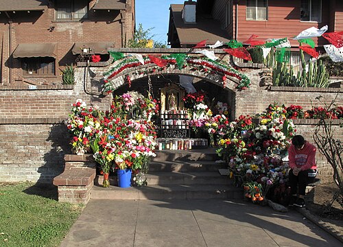 A street shrine to the Virgin of Guadalupe built into a brick archway on Kingsley Drive, banked with flowers and paper bunting.