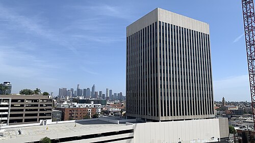 A high-rise office tower with the downtown Los Angeles skyline behind it, seen from a nearby rooftop parking deck.