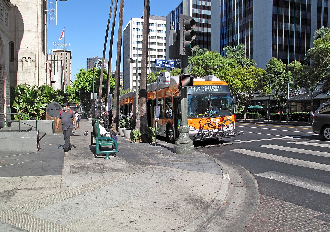 Wilshire Boulevard at Normandie Avenue, looking east past a Metro bus and palm trees toward downtown high-rises, with the Gaylord building's rooftop sign visible in the distance.