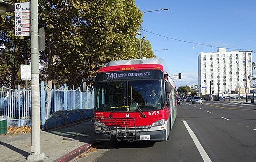 A Metro Rapid bus stopped at Vernon Avenue and Crenshaw Boulevard, a blue park fence and an apartment tower behind it.
