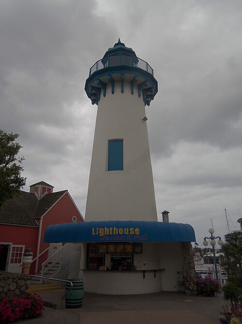 The white and blue lighthouse tower over the Lighthouse Fountain and Grill snack stand at Fisherman's Village, a red shingled roof beside it.