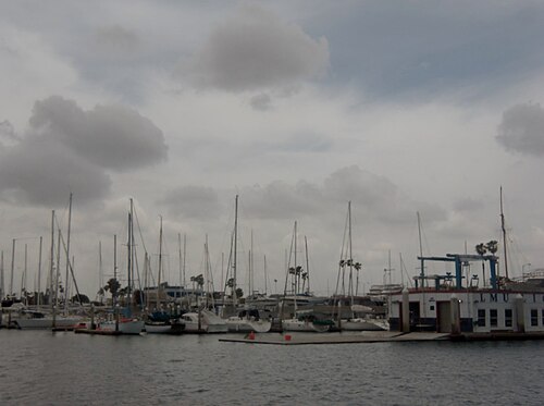 The LMU Lions boathouse, a white dockside building with a boat lift crane, moored sailboats lined up along its dock.