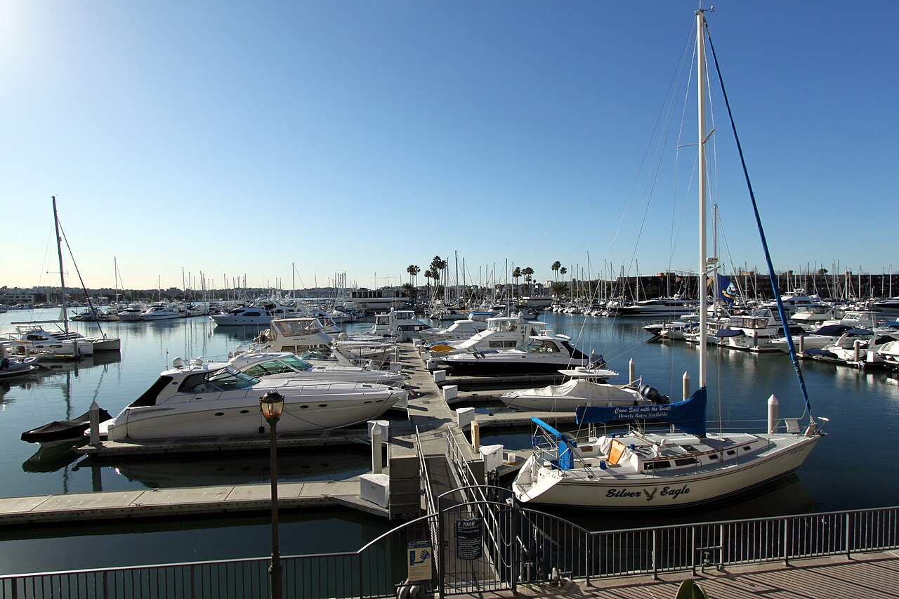 A floating dock gangway lined with moored sailboats and motor yachts, apartment buildings and palm trees along the basin's far shore.