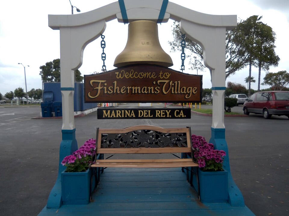 The wooden entrance arch and bell reading Welcome to Fisherman's Village, Marina Del Rey, over a bench in the parking lot off Fiji Way.