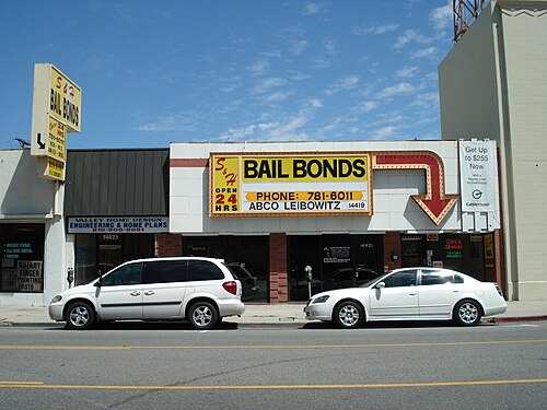 A row of storefronts on a Van Nuys street including S&H Bail Bonds, with an arrow shaped sign and cars parked at the curb.