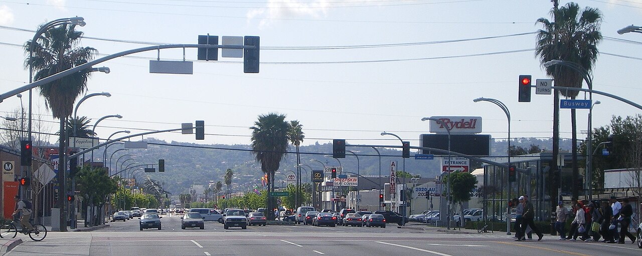 Van Nuys Boulevard looking north at a signalized intersection, palm trees and a Rydell car dealership sign on the right, low hills in the distance.
