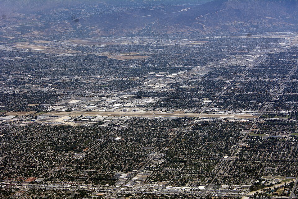 A wide aerial view of Van Nuys Airport and the surrounding San Fernando Valley, the runway running through blocks of hangars and warehouses with mountains behind.