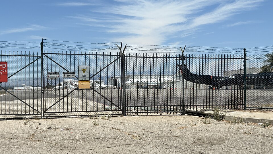 A locked gate in the perimeter fence at Van Nuys Airport, private jets parked on the ramp behind it.