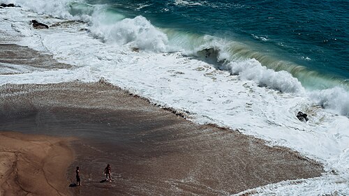A wave breaking across the sand seen from above, two people standing well back from the water.
