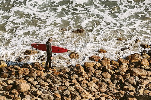 A surfer carrying a red board over a rocky stretch of shoreline, waves breaking behind him.