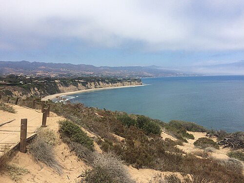 A sandy trail along the bluff top with a low wooden rail, the coastline and houses on the bluff visible beyond.
