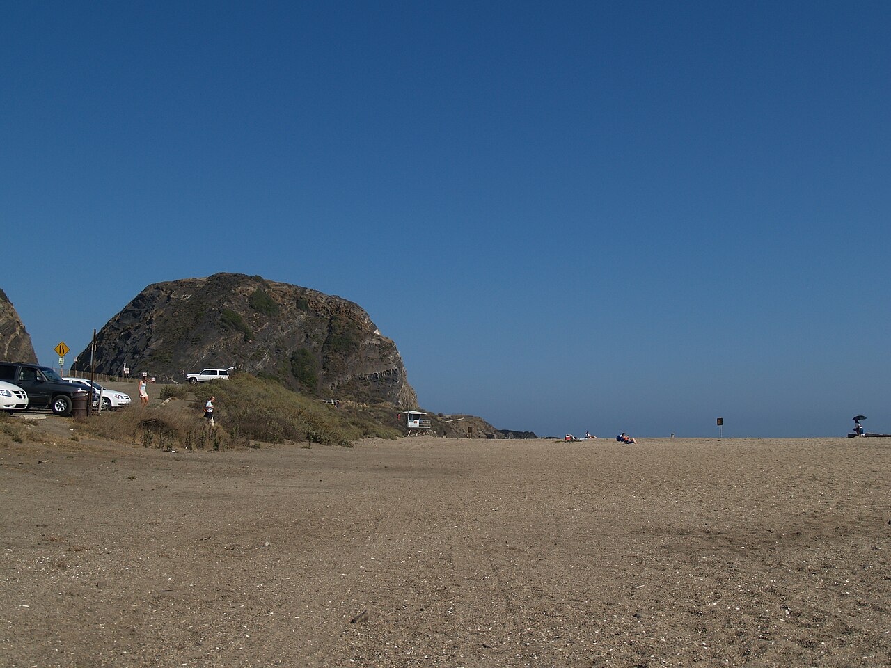 The beach at the foot of the headland on an ordinary sunny day, a few cars and people near the small parking area and the cliff rising behind them.