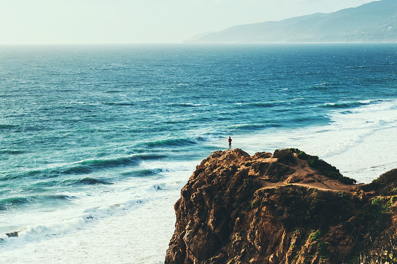 A person standing at the end of a rock outcrop above the water, waves breaking below and the coastline receding toward the mountains in the haze.