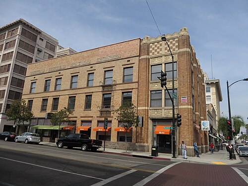 A tan brick corner building at Raymond Avenue and Colorado Boulevard, exterior fire escapes down one wall and a checkerboard brick parapet.