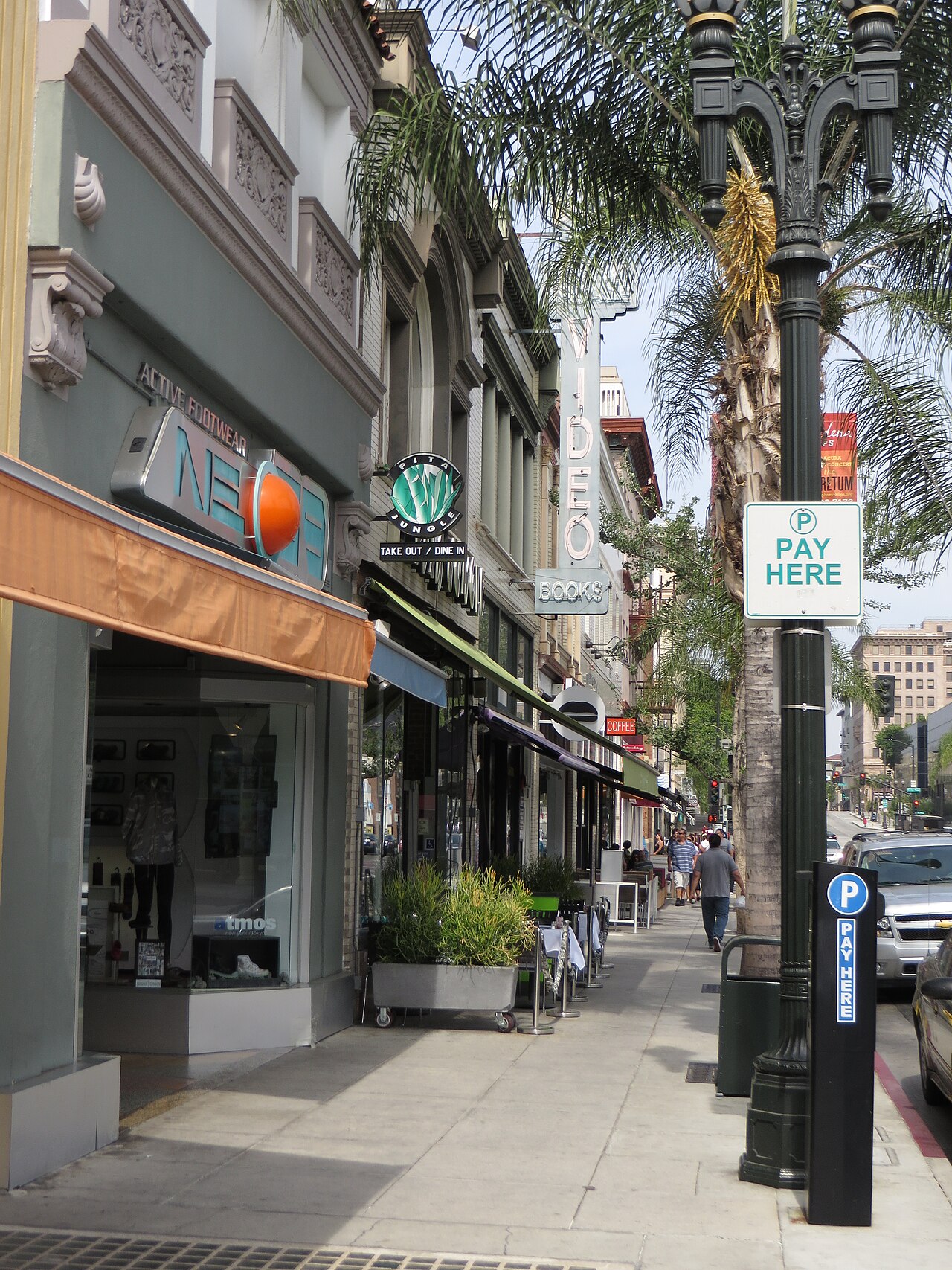 A sidewalk on Colorado Boulevard lined with storefront signs, ornate cornices above the shopfronts and a palm tree growing through the awnings.