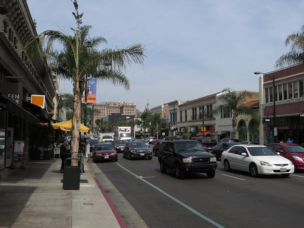 Colorado Boulevard on a weekday afternoon, a valet stand and café tables on the sidewalk, palm trees down the block and taller buildings visible in the distance.