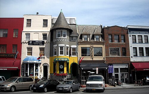 A row of shopfronts on Eighteenth Street under a grey conical turret and stepped gables, printed shop signs stacked over a narrow entrance, cars parked at the curb.